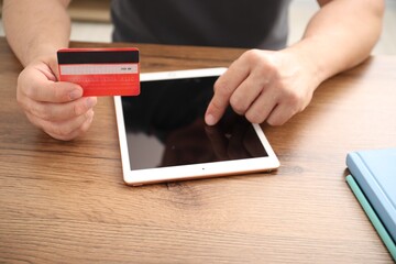 Man with credit card using tablet at wooden table indoors, closeup