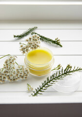 Balm made with Achillea millefolium, yarrow or common yarrow. Jar with medicinal balm on white wood background. Fresh flower for decoration. Still life. © FotoHelin