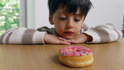 A young boy sitting at a table, hands clasped, intently gazing at a colorful sprinkled donut,...