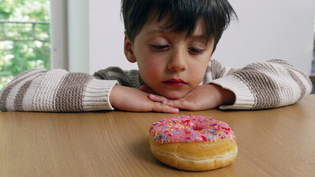 A young boy sitting at a table, hands clasped, intently gazing at a colorful sprinkled donut, demonstrating delayed gratification with a thoughtful and curious expression