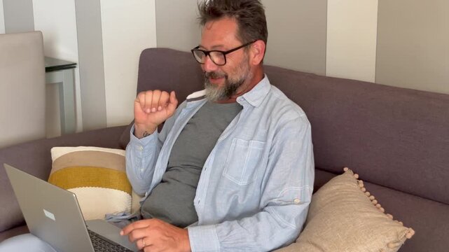 Carefree adult man sitting on the sofa talking in videocall using laptop and online wireless connection. Modern single male 50 yo speaking on computer call for business or friendship wearing glasses
