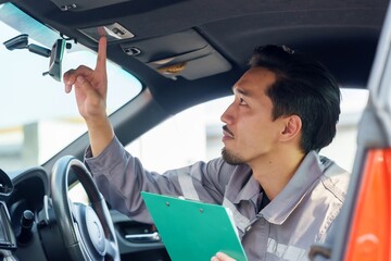 Asian male insurance officer in uniform using clipboard to inspect car interior. Pointing towards car roof light, focusing on thorough inspection during claim process. Represents attention to detail,