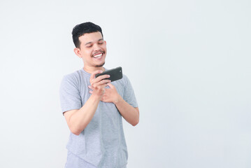 Portrait of an Indonesian Asian man, wearing a grey T-shirt, expressively playing a game on his smartphone, isolated against a white background.