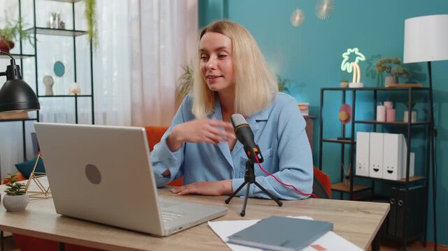 Happy blogger woman recording podcast at desk, giving workshop, webinar. Young Caucasian girl speaking at microphone and laptop webcam, making video call, discussing at home office in living room.