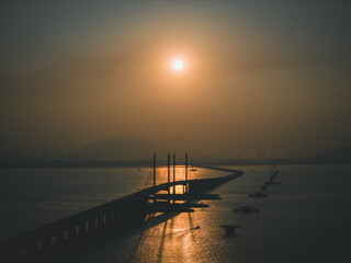 A stunning view of Penang Bridge at sunrise, framed by two TNB monopole electric transmission towers. The golden morning light casts a warm glow over the scene, highlighting the bridge's structure.