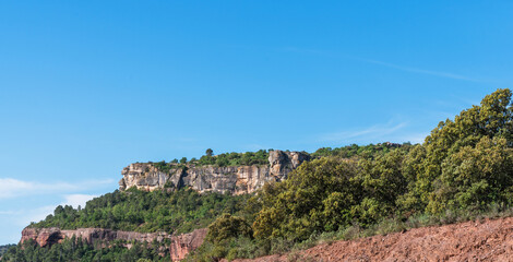 A serene landscape unfolds with a rugged, reddish-brown earth foreground leading to lush greenery, crowned by towering cliffs under a clear blue sky.