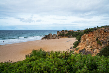 A secluded sandy beach bordered by rugged cliffs and Mediterranean shrubbery, with a solitary figure walking by the water under a partly cloudy sky.