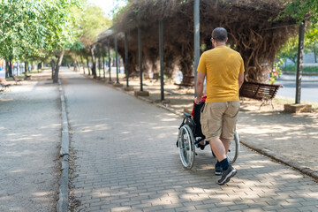 Obraz premium Man on his back pushing a wheelchair with an elderly woman sitting in a public park outside.