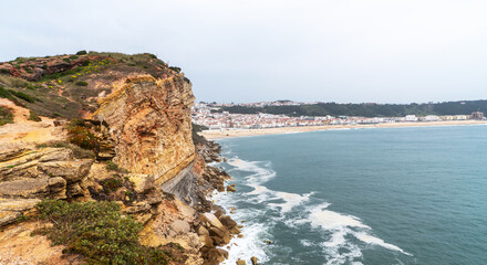 a stunning coastal view from atop a rugged cliff, overlooking a long stretch of sandy beach that reaches towards a quaint seaside town, under a muted sky.
