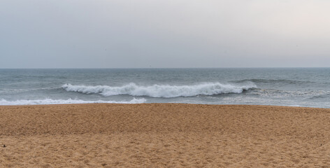 Overlooking a quiet beach with golden sands that meet rolling waves under a somber sky. Powerful waves cresting towards the sandy shore offering a sense of calm and solitude.