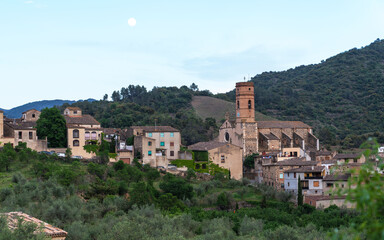 Fototapeta premium Spanish village nestled in a valley at dusk, with the moon hanging low in the sky. Traditional stone buildings with terracotta roofs line the foreground, backed by lush green hills.