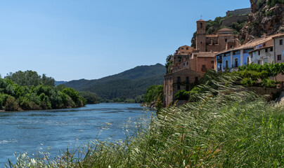 A serene river gently flows by a historic village with terracotta roofs, nestled against a backdrop of lush hills under a clear blue sky, capturing the essence of rural tranquility