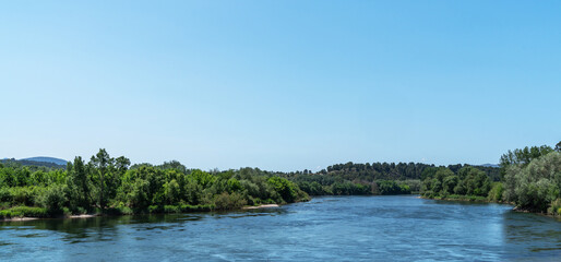 A tranquil river cuts through lush foliage under an expansive blue sky, with distant hills rolling into the horizon, embodying serene natural beauty.