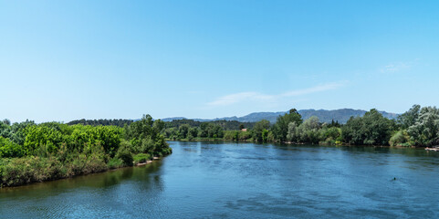 Overlooking a serene river, the foreground showcases a rustic terracotta-tiled roof, with lush greenery on the banks and distant mountains under a clear expansive sky.