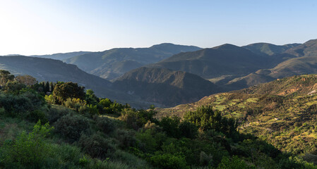 Early morning light bathes a serene landscape, highlighting the lush greenery of rolling hills and valleys, with layers of mountains receding into the distance under a clear sky