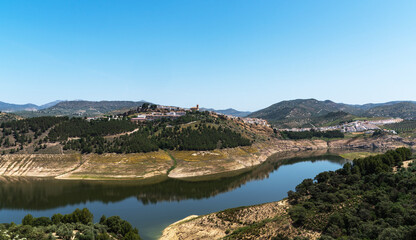 Fototapeta premium a serene lake nestled in a valley, with a quaint town perched atop a hill surrounded by a patchwork of olive groves under a cloudless blue sky