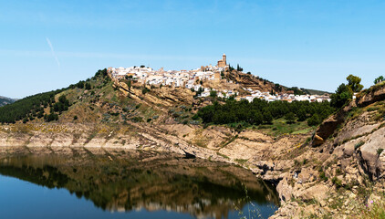 Fototapeta premium picturesque landscape a hilltop Spanish village with clustered white houses and a church, reflected in the tranquil waters of a foreground reservoir, a backdrop of clear blue skies and hills.
