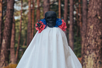 Ghostly bride figure draped in white with black skull mask adorned with blue and red flowers stands...