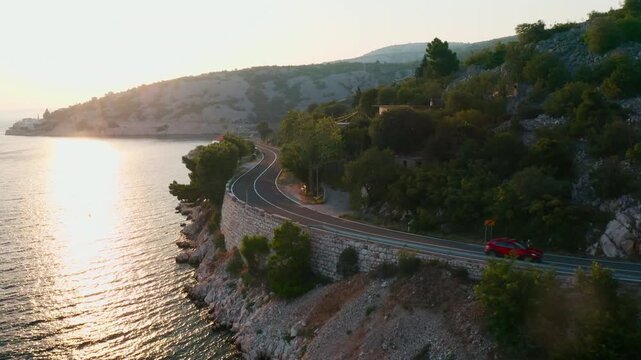 Passenger car passing on double-lane highway near sea reflecting sunlight. Route at slope foot near water with mountains on background aerial view