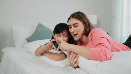 Happy asian daughter and smart mom lie on bed while looking at camera. Energetic family spend time and prepare to sleep together. Cute girl and mom smile to camera show love and affection. Pedagogy.