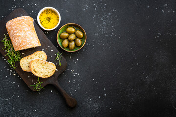 Ciabatta bread on wooden board with olive oil, olives and herbs on black. Top view with copy space.