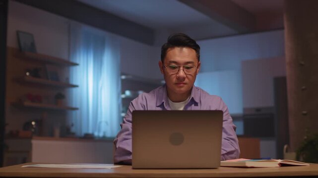 Serious asian businessman with glasses use laptop typing on keyboard and make notes. Working at night time. 360 degree. Camera moving around