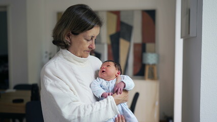 Grandmother holding newborn baby grandson in arms, first week of life of infant, standing in living-room