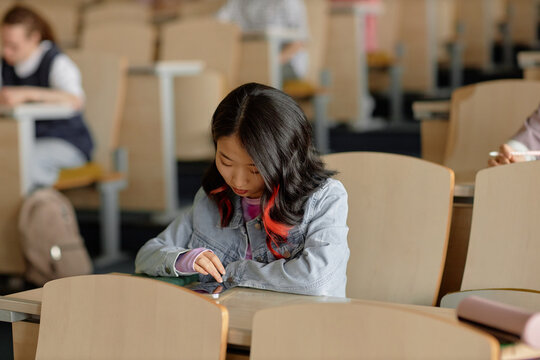Young Asian female student on phone checking social media and chatting during university lecture, copy space