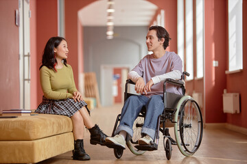 Two young multiethnic students chatting away amicably in university hallway. Smiling young man with disability talking to Asian girl, copy space