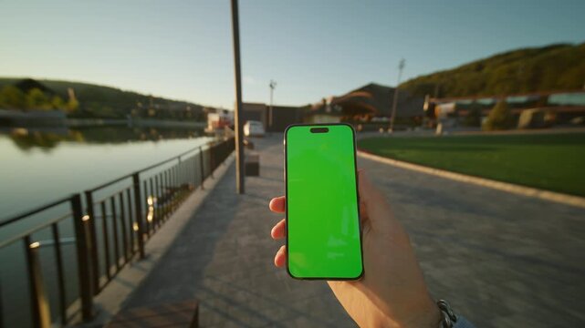 Hands man holding vertically phone with green screen. On background lake on sunlight. Nature. Smartphone device