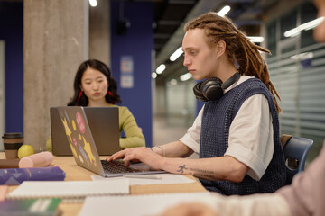 Side view of young nonconforming dreadlocked male student practicing writing skills studying on laptop while sitting at large desk at university group seminar