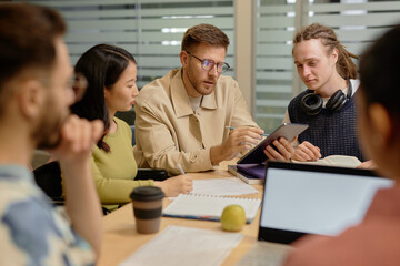 Adult male teacher explaining task to multiethnic college students pointing at digital tablet...