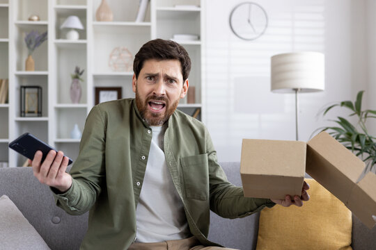 Frustrated man holding smartphone and open delivery box expressing disappointment. Emotional reaction to unsatisfactory online shopping experience, customer dissatisfaction delivery problem at home.