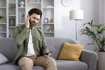 Man sitting on couch experiencing ear pain while holding smartphone in hand. He looks uncomfortable and in pain. Home setting with modern decor visible in background.