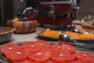 Close-up of a plate with red grapefruit slices in a high-end hotel dining area. Background includes assorted fruits and a red meat slicer, showcasing luxury and detail.