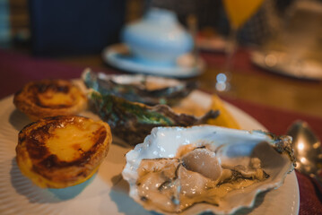 Close-up of a plate featuring fresh oysters and custard tarts in a refined table setting with a blue teapot and orange juice, suggesting a high-end restaurant.