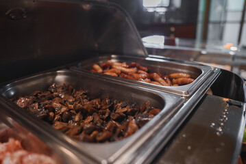 Close-up of a luxury hotel's buffet featuring sauteed mushrooms and assorted sausages in stainless steel chafing dishes. Sophisticated interior with ambient lighting.
