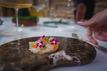 A beautifully presented seafood dish with edible flowers on a dark marble-patterned plate. The elegant table setting and manicured hand suggest a high-end dining experience.