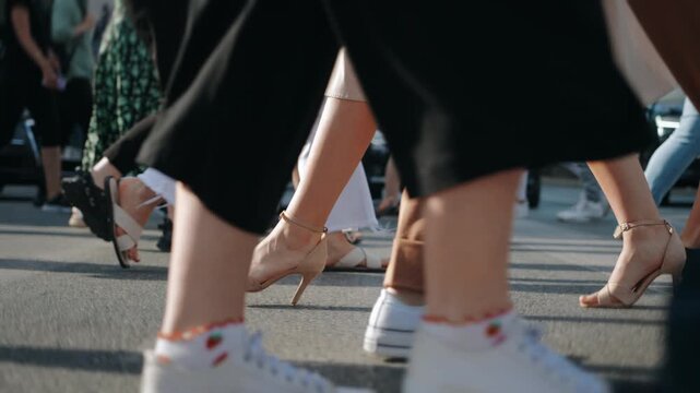 Following female feet steps crossing busy street. Slim woman wearing dress and high heels goes on pedestrian crossing in city. Low angle people crowd legs walking. No visible brand names and logos