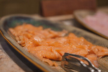 Beautifully presented thinly sliced salmon on a ceramic platter with metal tongs, part of a luxury hotel's gourmet dining experience. Background slightly blurred.
