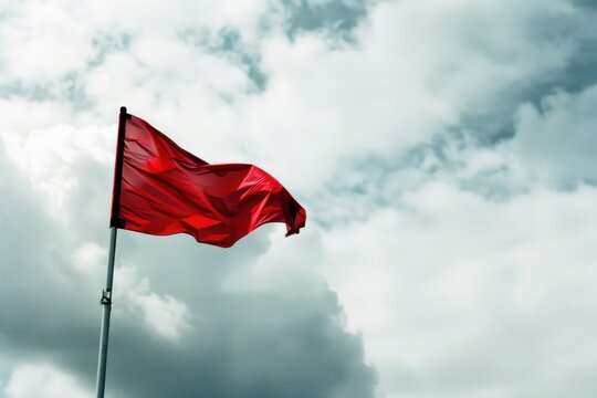 A red flag waves prominently against a cloudy sky, symbolizing warning, alertness, or danger, with dramatic and bold color contrast.