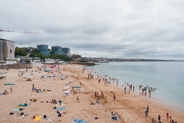A vibrant coastal city beach with people enjoying the sandy shore, colorful umbrellas, and calm waters. Modern high-rise buildings and a historic castle-like structure in the background.