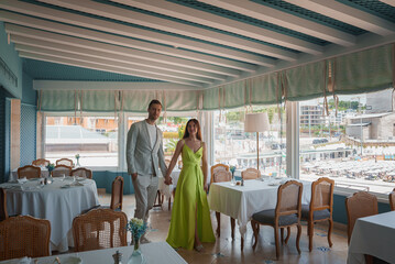 A well-dressed couple in a beautifully designed dining area of a luxury hotel. Features elegant decor, light blue walls, and large windows with a beach view.