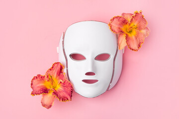 Cosmetic LED facial mask regenerative treatment on pink background with flowers lilies. Health and...
