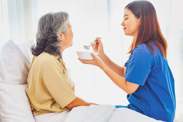 Sick elderly woman is closely cared for female nurse who is taking care of her. She slowly scoops...