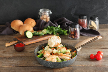 Frying pan with tasty chicken fillet, vegetables, buns and spices on wooden table