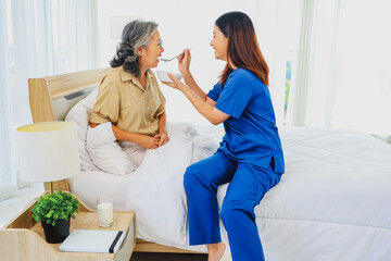 Obraz premium Sick elderly woman is closely cared for female nurse who is taking care of her. She slowly scoops porridge into her mouth nutritious cooked breakfast. Help her recover from her illness in bed at home.