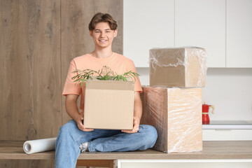 Young man with cardboard boxes and roll of stretch film sitting on table in kitchen