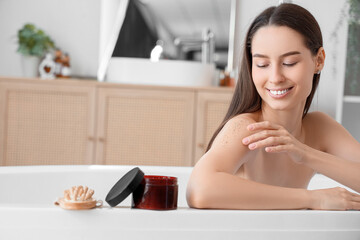 Obraz premium Beautiful young happy woman with jar of body scrub and massage brush sitting in bathroom