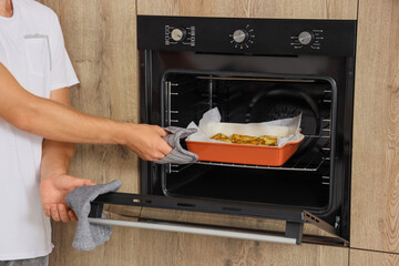 Young man taking baking dish with potato from oven in kitchen, closeup